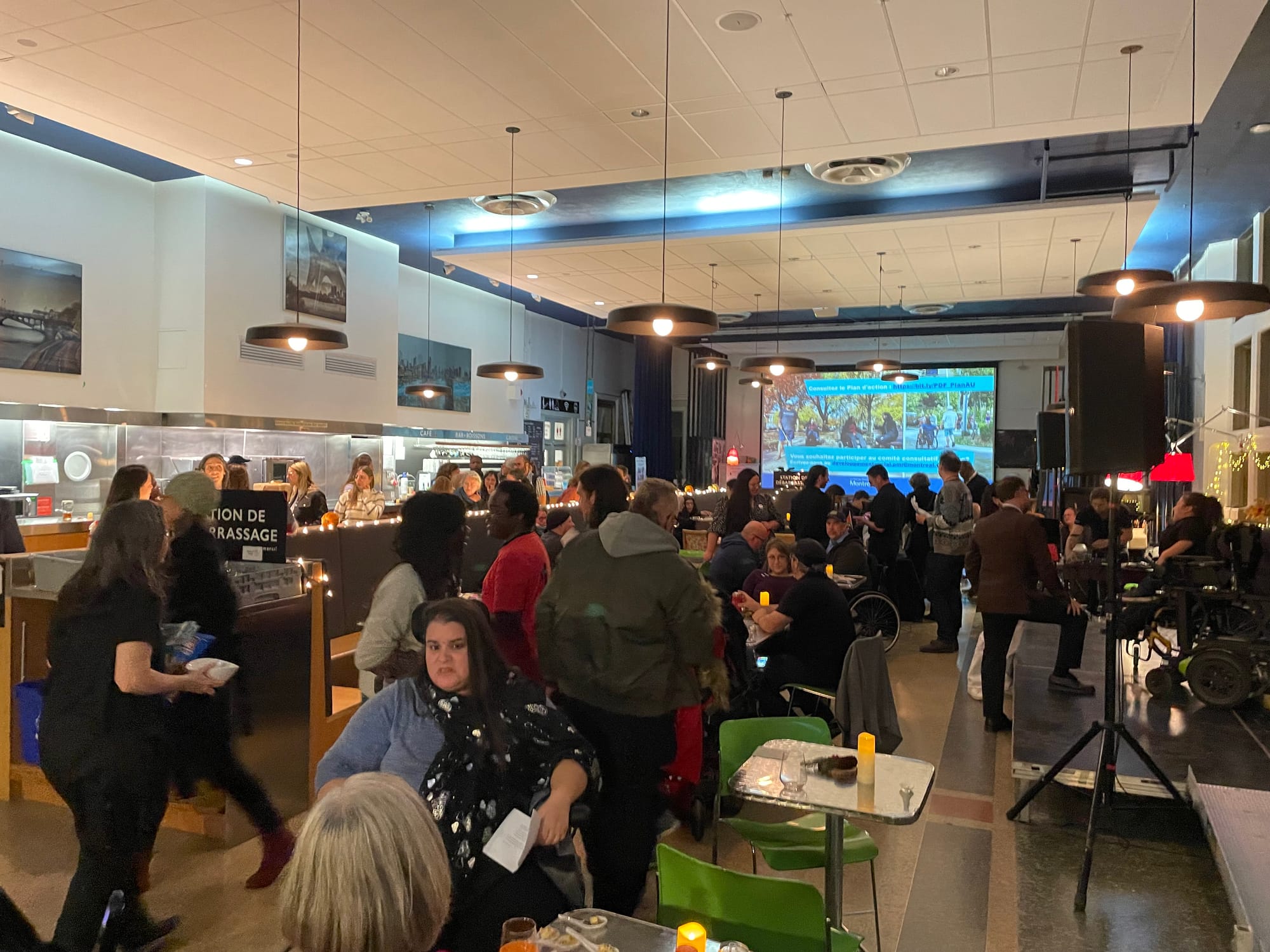 Cette photo montre une foule majoritairement assise dans une grande salle avec des tables où les gens discutent entre eux.