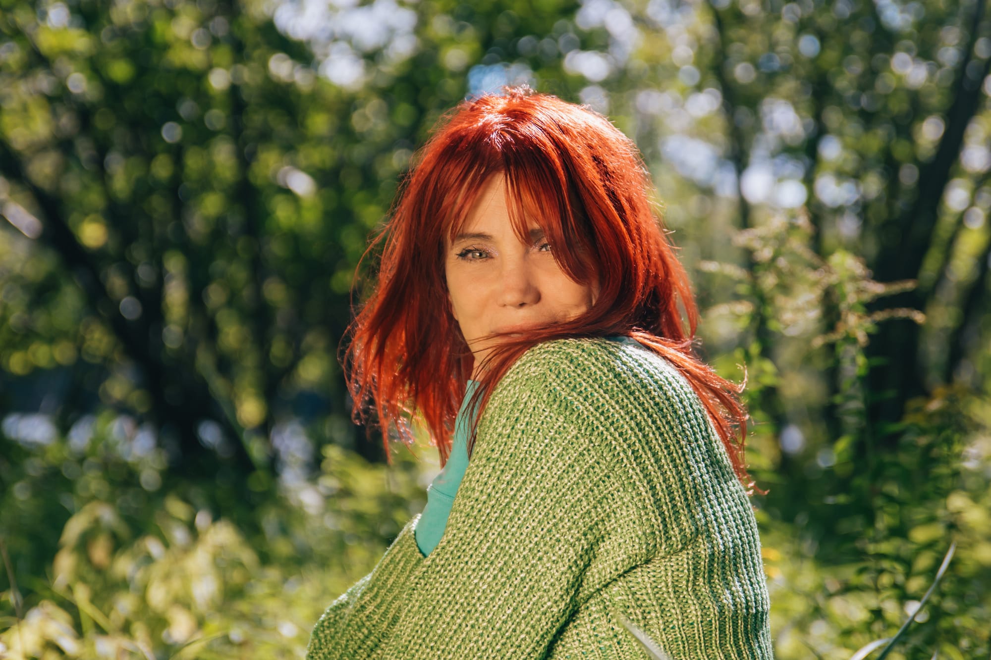 Une femme aux cheveux rouges, avec un chandail vert, dehors devant des arbres. On devine un cours d'eau à l'arrière.