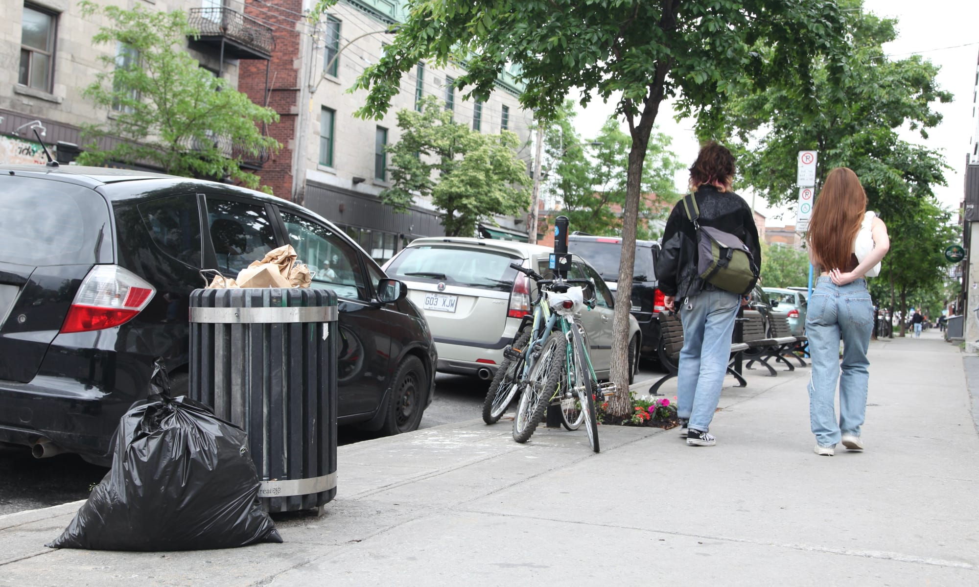 Un sac de déchets repose aux côté d'une poubelle de la rue Saint-Viateur, alors que deux femmes marches sur le trottoir un peu plus loin, dépassant des vélos stationnés près d'un arbre.