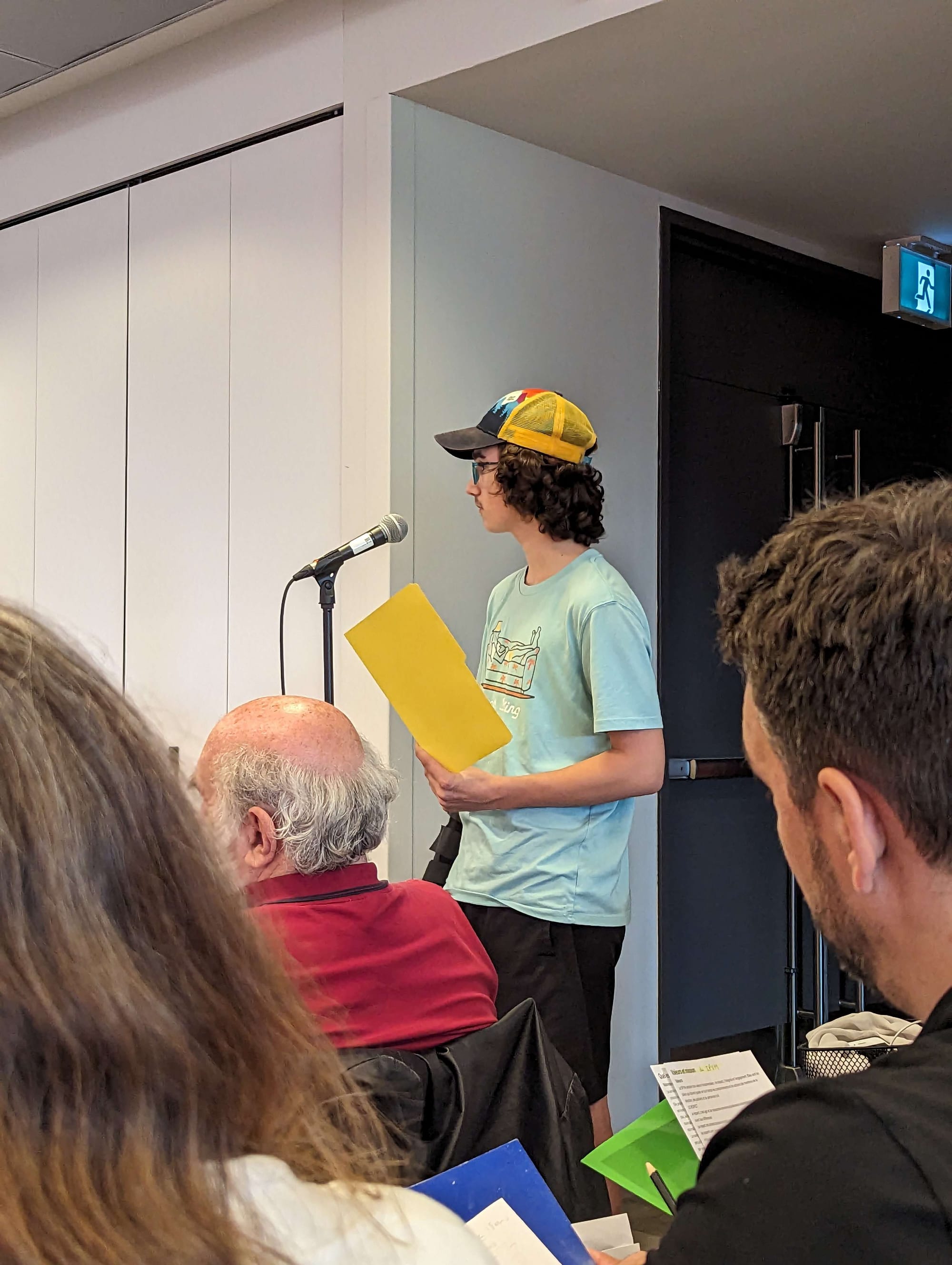 Un jeune homme debout devant quelques membres du public assis, attend son tour de parole devant un microphone, alors qu'il tient un dossier jaune dans ses mains. 