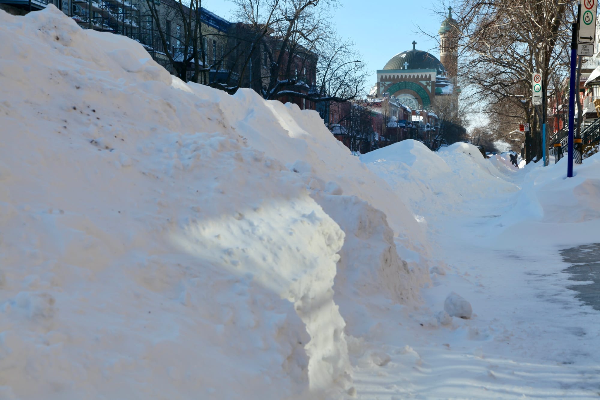 On voit un tas de neige de plus de 1,5m le long d’un trottoir.